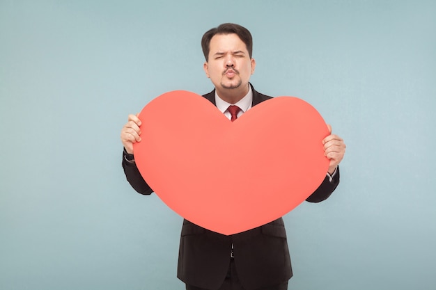 Con amor, st. concepto de San Valentín. Hombre de negocios con gran corazón, enviar beso. Interior, foto de estudio, aislado sobre fondo azul claro o gris