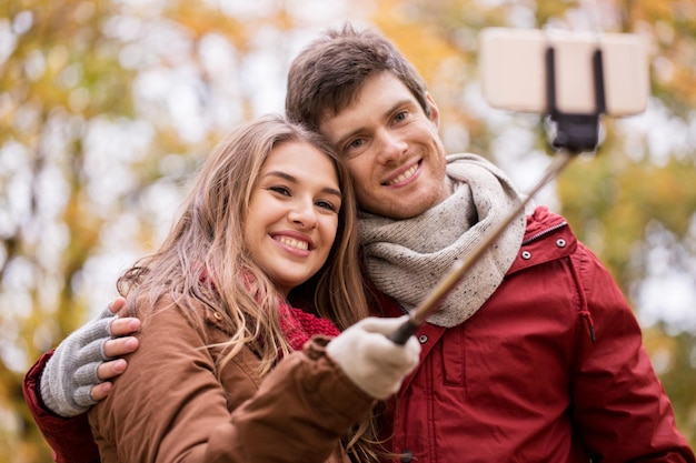 amor, tecnologia, relacionamento, família e conceito de pessoas - casal feliz e sorridente tirando fotos com bastão de selfie de smartphone no parque de outono