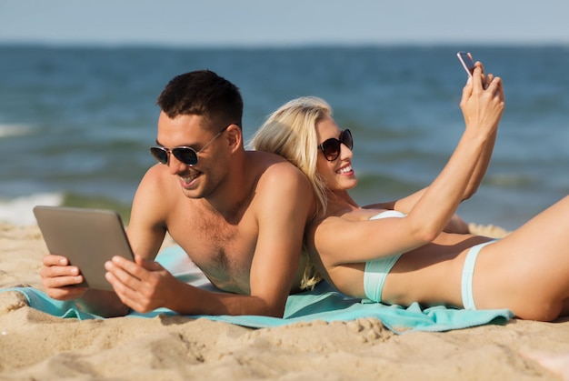 amor, viajes, turismo, tecnología y concepto de personas - pareja feliz de vacaciones con tablet pc y smartphone tomando el sol en la playa de verano