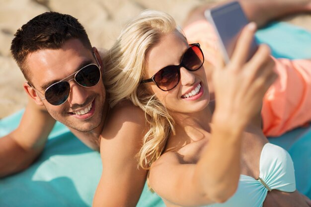 amor, viajes, turismo, tecnología y concepto de personas - pareja sonriente de vacaciones en traje de baño y gafas de sol y tomando selfie con smartphone en la playa de verano