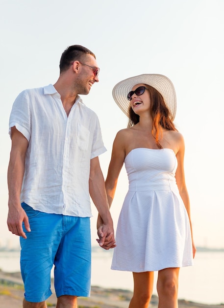 el amor, los viajes, el turismo, el verano y el concepto de la gente - pareja sonriente de vacaciones con gafas de sol y tomándose de la mano caminando por la playa