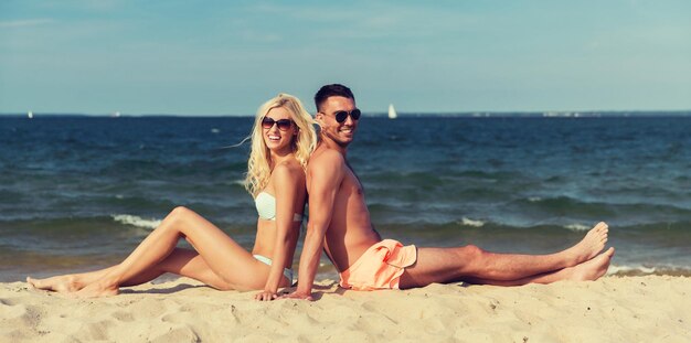el amor, los viajes, el turismo, el verano y el concepto de la gente - pareja sonriente de vacaciones en traje de baño sentado en la playa espalda con espalda
