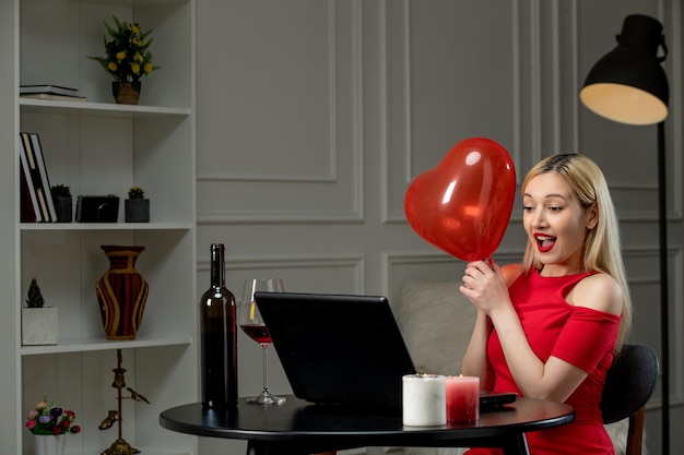 Amor virtual linda chica rubia con vestido rojo en una cita a distancia con vino emocionada por el globo del corazón