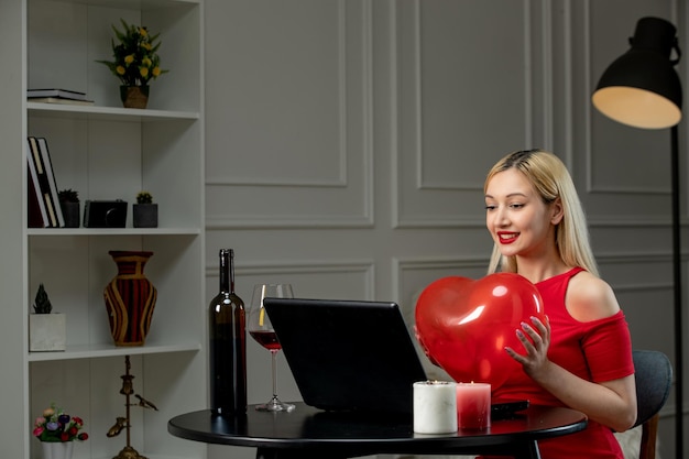 Amor virtual linda chica rubia en vestido rojo en cita a distancia con vino sosteniendo globo de corazón