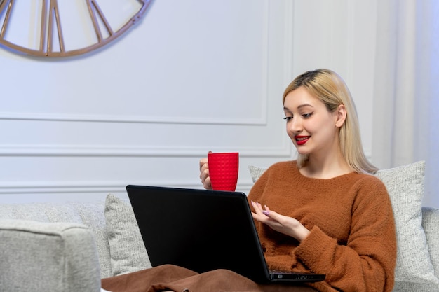 Amor virtual linda jovencita rubia en suéter acogedor en fecha de distancia de computadora sosteniendo taza roja