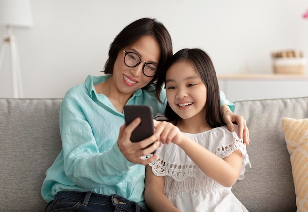 Amorosa abuela asiática y su linda nieta usando un teléfono inteligente en el sofá en casa