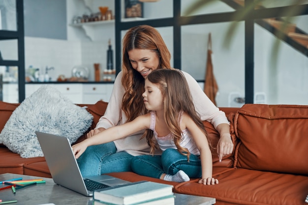 Amorosa e linda mãe e sua filha se unindo e sorrindo enquanto usam o laptop em casa