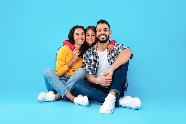 Amorosa familia árabe abrazando sentado en estudio sobre fondo azul.