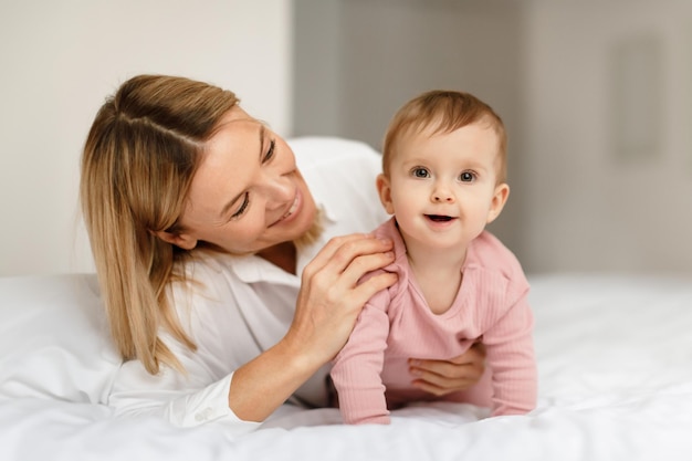 Amorosa madre hablando y jugando con su bebé niña niño gateando y sonriendo a la cámara en la cama