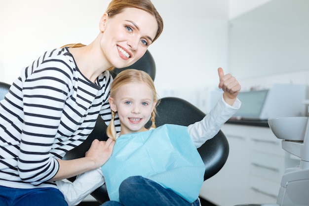 Amorosa madre joven abrazando a su pequeña hija y posando junto con ella mientras la niña sentada en la silla del dentista y esperando un chequeo