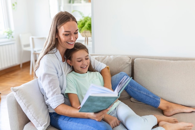 Amorosa madre joven leyendo un libro a su adorable hijita, sentada en un acogedor sofá en la sala de estar, mamá enseñando a una niña en edad preescolar, familia pasando el fin de semana en casa juntos, educación infantil