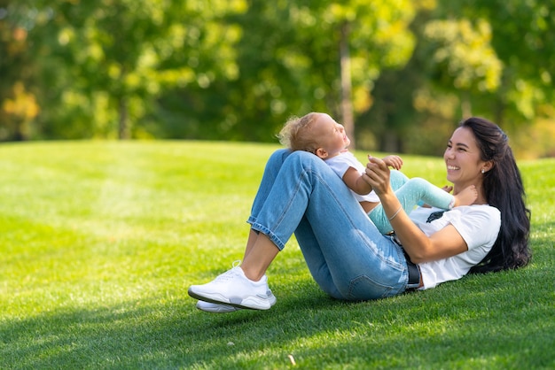 Amorosa madre joven riendo con su bebé mientras juegan juntos en la hierba verde en un parque o jardín a la sombra de un árbol