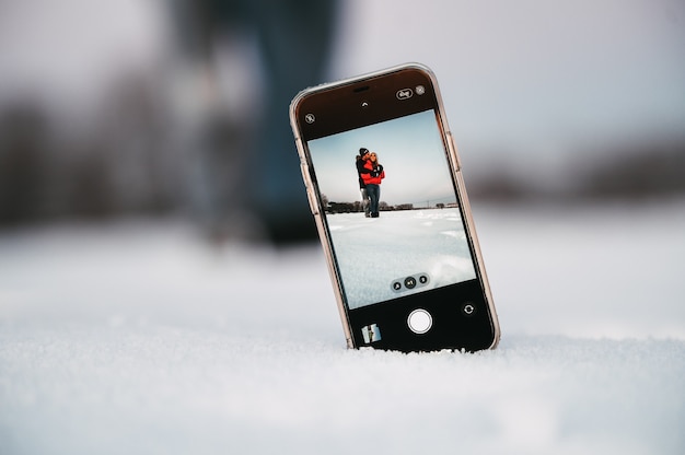 Amorosa pareja abrazándose mientras toma selfie en teléfono móvil colocado sobre la nieve en el campo