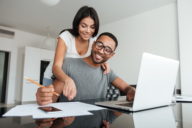 Amorosa pareja joven usando laptop y analizando sus finanzas. Escribir notas.