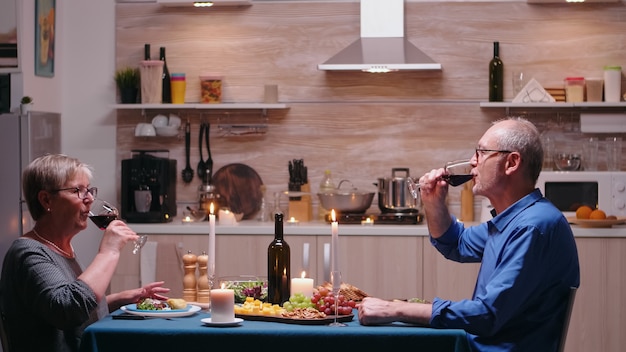 Amorosa pareja madura levantando copa de champán y brindando mientras disfruta de una cena romántica en casa en la cocina. Ancianos mayores comiendo la comida, celebrando su aniversario en el comedor.