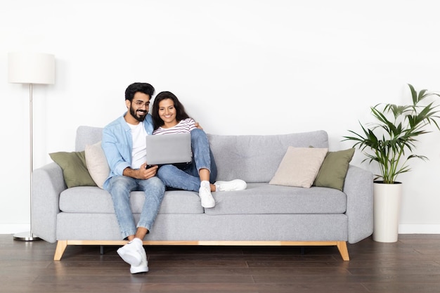 Foto amorosa pareja oriental sentada en el sofá en casa usando una computadora portátil