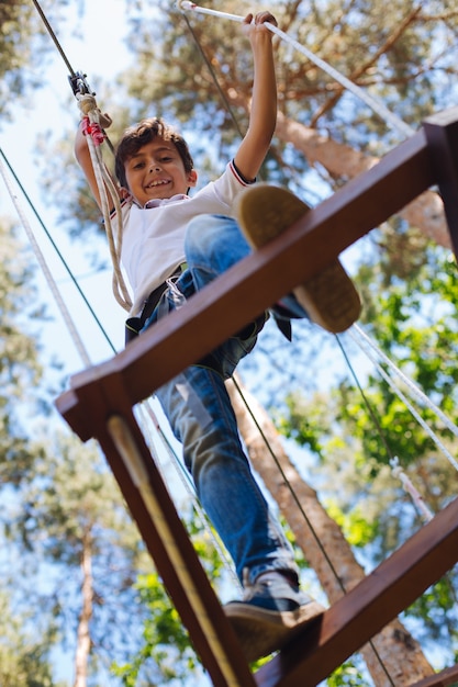 Amoroso extremo. Alegre niño preadolescente mirando hacia abajo y sonriendo a la cámara mientras da un paso al siguiente bar en un parque de cuerdas