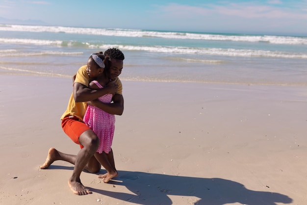 Amoroso joven afroamericano abrazando a su hija en la playa contra el mar durante el día soleado