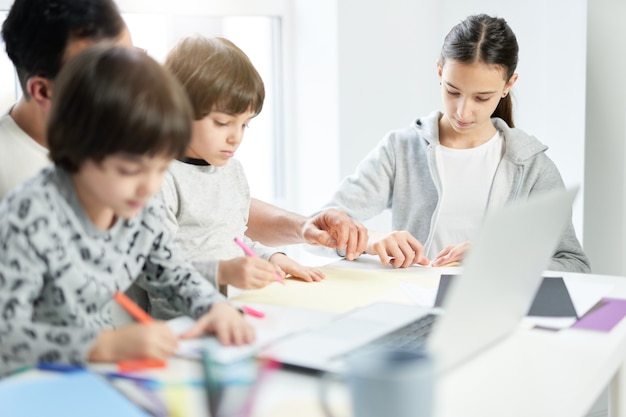 Amoroso padre latino jugando con sus hijos. Hombre de negocios que usa la computadora portátil mientras trabaja desde casa y mira a los niños. Freelance, encierro, concepto de familia