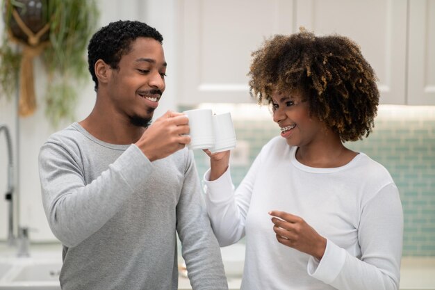 Amorosos cônjuges negros torcendo com canecas de café interior da cozinha