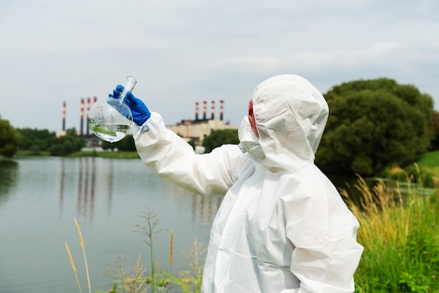 Amostragem de águas abertas. um cientista ou biólogo coleta uma amostra de água perto de uma planta industrial. uma amostra de água em um frasco químico redondo.