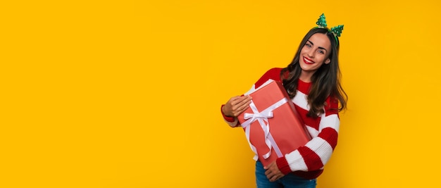 Amplia foto de banner de hermosa mujer sonriente emocionada con caja de regalo de Navidad en las manos se divierte mientras posa sobre fondo amarillo