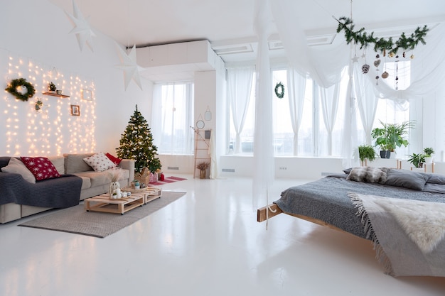 Amplio y luminoso apartamento de estilo balinés de planta abierta, espacioso, con cama colgante y ventanas panorámicas. piso y paredes blancos, muebles sencillos de madera. decorado con un árbol de navidad