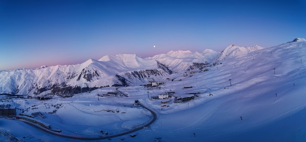 Amplio panorama aéreo de la cordillera nevada en el amanecer de invierno Impresionante cordillera cubierta de nieve en polvo en la estación de esquí al atardecer Luna sobre el horizonte de los picos del Cáucaso en el atardecer por la noche