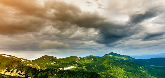 Amplio panorama de iluminado por el sol de la mañana verde valle, colinas cubiertas de bosque y distantes montañas brumosas con parches de nieve bajo el cielo nublado y ventoso. Belleza de la naturaleza, el turismo y el concepto de viaje.