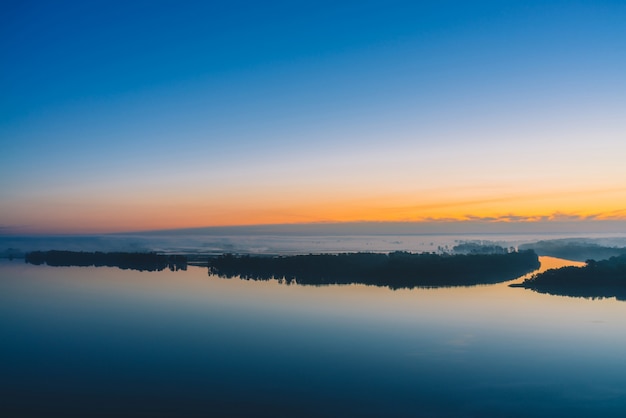 Amplio río fluye a lo largo de la orilla diagonal con bosque bajo una espesa niebla. Principios del cielo azul se refleja en el agua. Resplandor amarillo en el pintoresco cielo antes del amanecer.