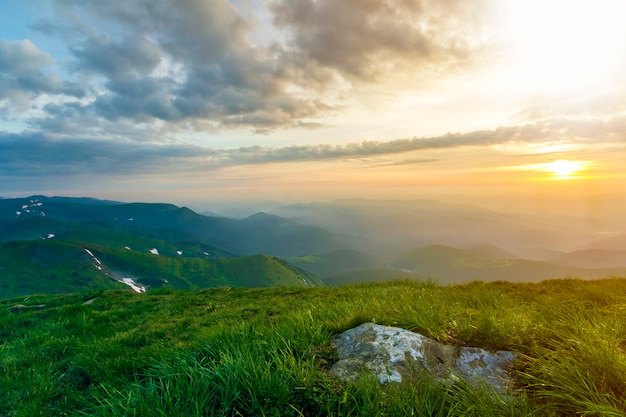 Amplio verano vista a la montaña al amanecer. Sol naranja brillante en el azul cielo nublado sobre la colina cubierta de hierba verde con gran roca y cordillera distante cubierto de niebla de la mañana. Concepto de belleza de la naturaleza.