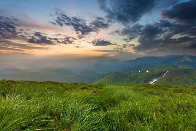 Amplio verano vista a la montaña al amanecer. Sol naranja brillante que se eleva en el azul cielo nublado sobre la colina cubierta de hierba verde hierba suave y cordillera distante cubierto de niebla de la mañana. Belleza del concepto de naturaleza.