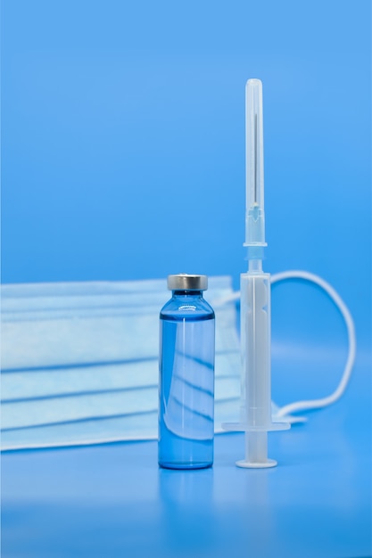 Ampollas médicas de vidrio, jeringa y máscara protectora sobre fondo azul. Productos farmacéuticos, vacunación