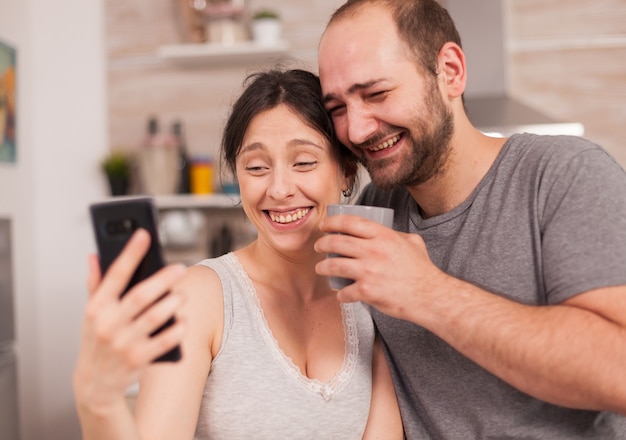 Amüsierte Paare, die morgens während des Frühstücks auf das Telefon schauen. Fröhlicher verheirateter Ehemann und Ehefrau, die beim Frühstück in der Küche lustige Gesichter machen.