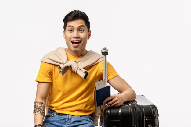 Amüsierter, fröhlicher, gutaussehender asiatischer junger männlicher Student reist schließlich ins Ausland, wartet im Flughafen-Boarding, sitzt mit schwarzem Koffer, gepacktem Gepäck und lächelt verträumt, weiße Wand