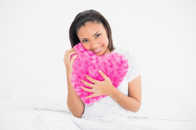 Amüsiertes junges dunkelhaariges Modell, das ein heartshaped Kissen streichelt