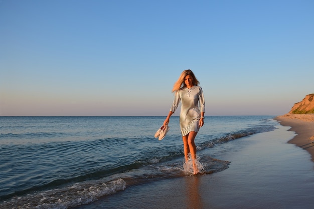 An der Küste vor blauem Himmel geht ein blondes Mädchen mit Schuhen in der Hand über das Wasser