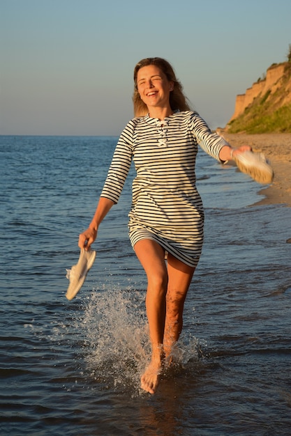 An der Küste vor dem Hintergrund des Himmels läuft ein blondes Mädchen auf dem Wasser, lächelt und hält Schuhe in der Hand