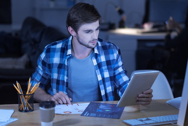 Análise de dados. homem bonito e inteligente que trabalha duro segurando o tablet e olhando seus documentos enquanto analisa os dados