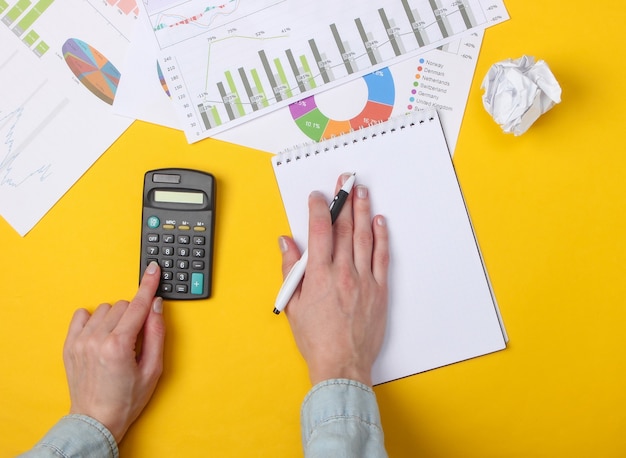 Análisis Economico. Manos femeninas cuentan la calculadora en un amarillo con un cuaderno, gráficos y tablas.