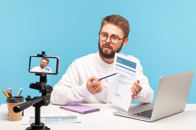 Analista de hombre barbudo concentrado explicando cómo trabajar con datos sentados en el lugar de trabajo en la oficina y posando en el tutorial de grabación de la cámara del teléfono inteligente Toma de estudio interior aislada en fondo azul