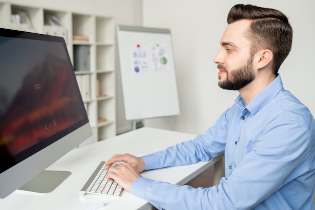 Foto analista joven ocupado sentado junto al escritorio frente a la pantalla de la computadora mientras escribe y busca información o sitios web