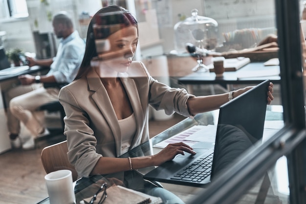 Analizando datos. Vista superior de la mujer joven moderna que usa la computadora mientras trabaja detrás de la pared de vidrio en la oficina