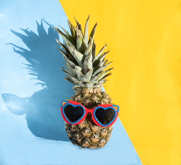 Ananas in herzförmiger Sonnenbrille auf halb blauem und halb gelbem Hintergrund