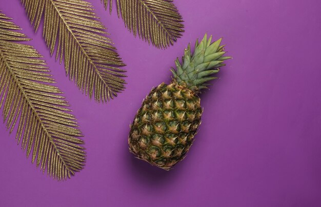 Ananas mit goldenen Palmblättern auf lila Hintergrund. Tropisches Konzept. Draufsicht