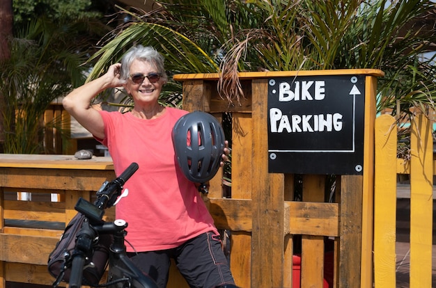 Anciana activa en excursión con su bicicleta con casco protector se detiene en el bar de aperitivos con zona de aparcamiento para ciclistas