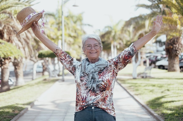 Una anciana alegre y atractiva parada en un parque público con los brazos extendidos sosteniendo su sombrero mirando a la cámara