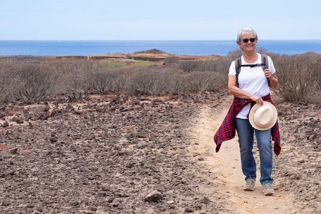 Una anciana alegre caminando por un sendero con mochila disfrutando de la excursión al aire libre Horizonte sobre el mar y el día soleado