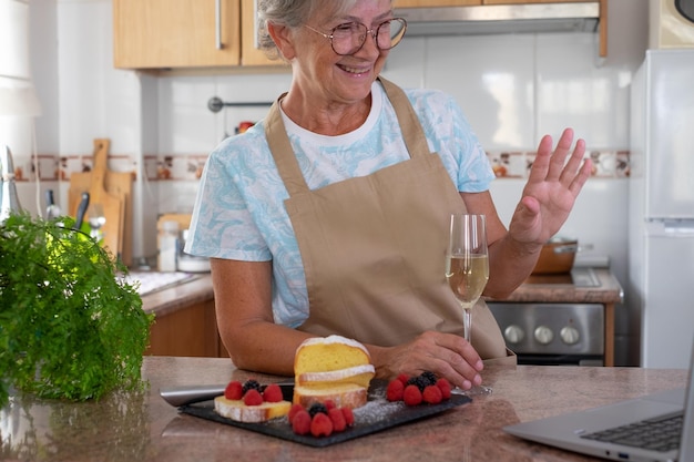 Una anciana alegre en la cocina de la casa después de hornear su tarta casera sosteniendo una copa de vino mientras conversaba con amigos o familiares por computadora portátil