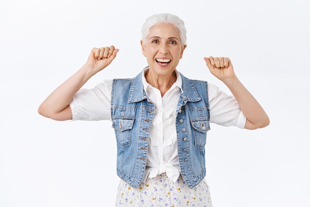 Una anciana alegre y despreocupada con el pelo gris apoya a su nieta durante la competencia levantando las manos triunfante sonriendo alegremente viendo el juego animando el fondo blanco